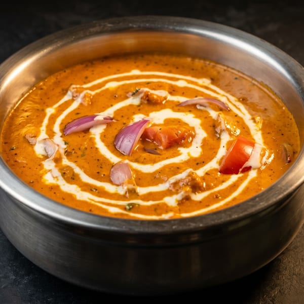 A metal bowl filled with creamy orange curry, garnished with white swirls, red onions, and tomatoes.