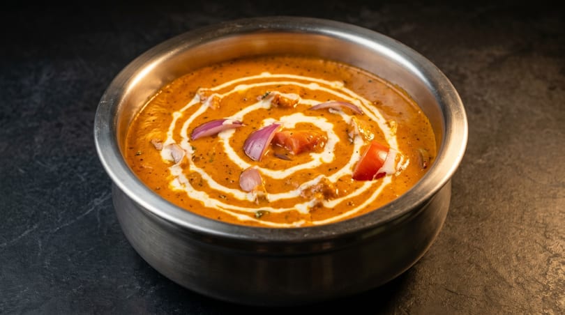 A metal bowl filled with creamy orange curry, garnished with white swirls, red onions, and tomatoes.