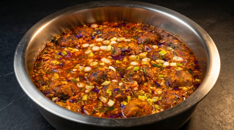 Close-up of a large metal bowl filled with Manchurian, garnished with chopped scallions and red cabbage.