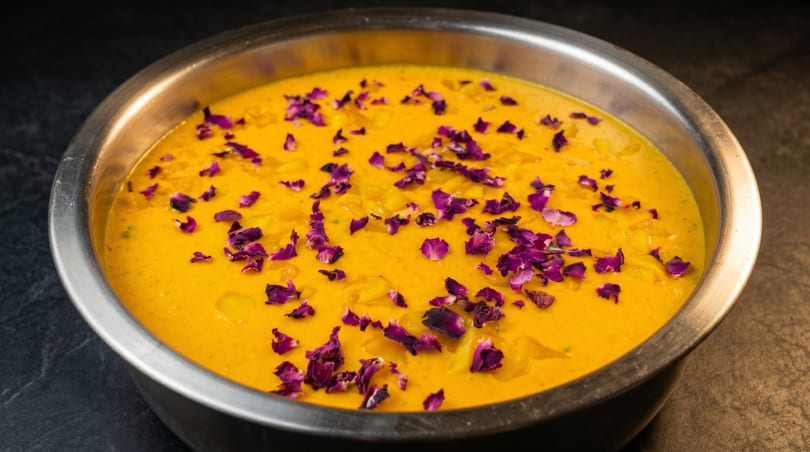 Golden mango curry with purple rose petals in a metal bowl on a dark surface.