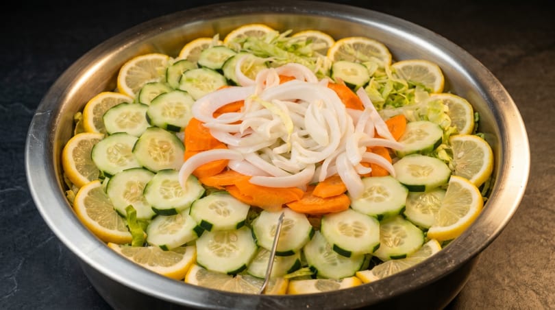 A large metal bowl filled with a fresh salad of sliced lemons, cucumbers, carrots, and onions.