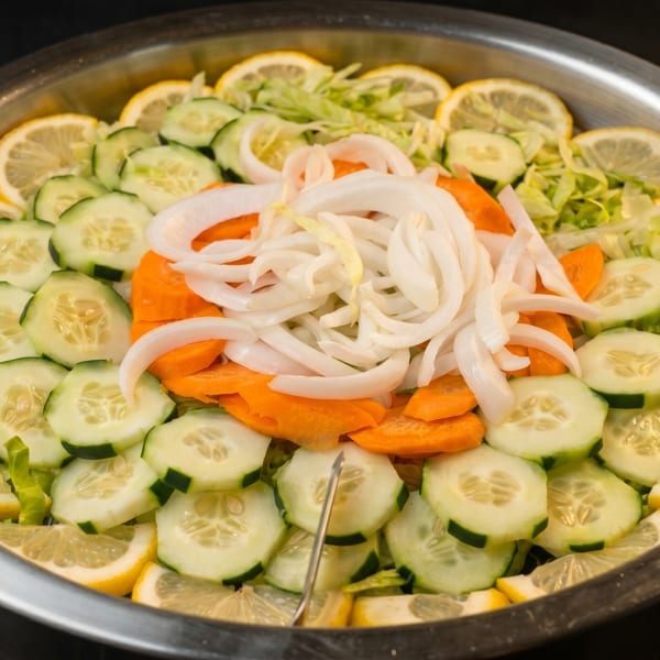 A large metal bowl filled with a fresh salad of sliced cucumbers, lemons, carrots, and onions.