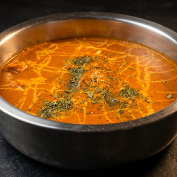 A metal bowl filled with orange curry, garnished with green herbs, on a dark textured surface.