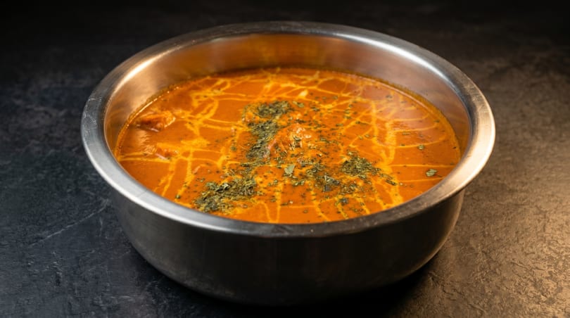A metal bowl filled with orange curry, garnished with green herbs, on a dark textured surface.
