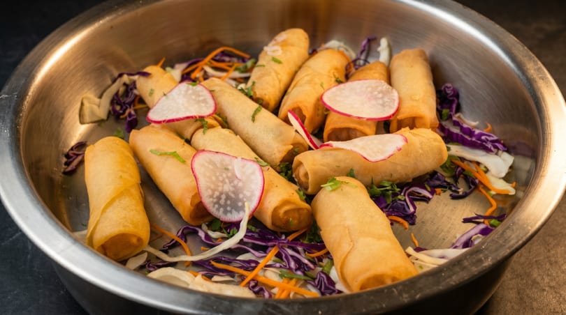 Golden spring rolls with radish slices and shredded cabbage in a metal serving dish.