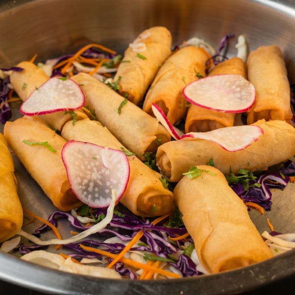 Golden spring rolls garnished with radish slices and shredded cabbage in a metal serving dish.